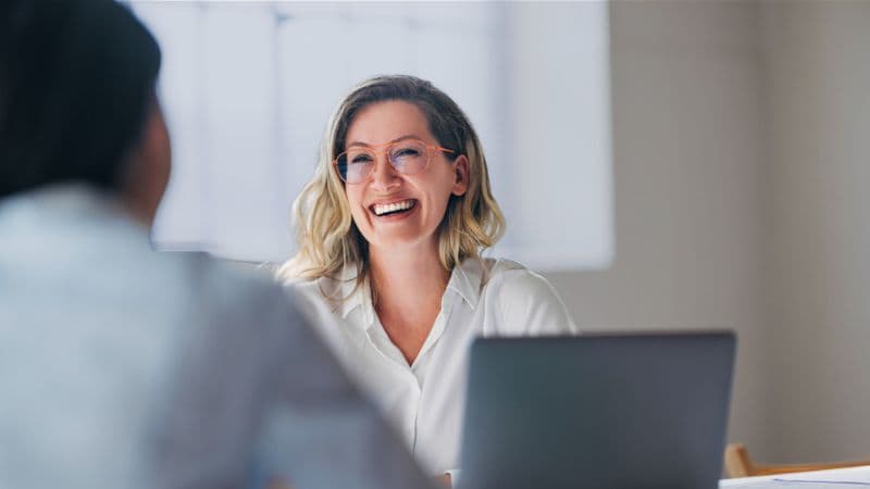 A Cheerful Woman With Glasses Is Sitting At A Desk, Engaged In A Interview Meeting